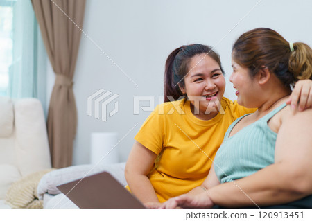 Happy Asian women friend relaxing in living room and drink a cup of tea together. Happy - delighted two Asian oversized women talking and watching a movie on laptop or notebook computer. Happy Asian women friend relaxing in living room and drink a cup of tea together. Happy - delighted two Asian oversized women talking and watching a movie on laptop or notebook computer. 120913451