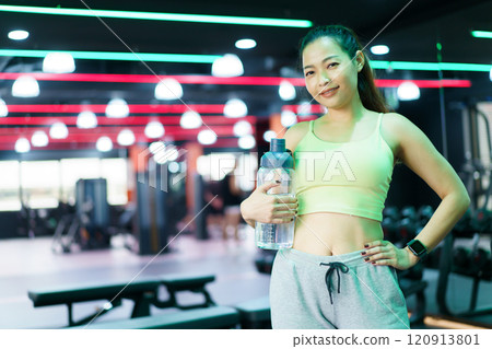 Good looking - beautiful Asian woman relaxing in the gym after made weight training and cardio workout, woman sitting or resting on the bench while drinking water after exercise. 120913801