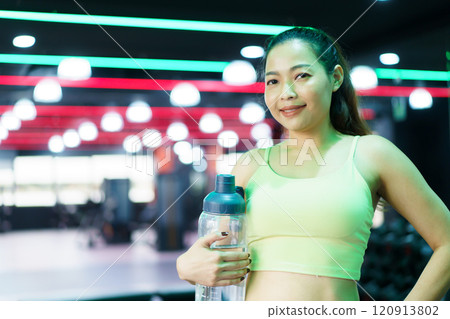 Good looking - beautiful Asian woman relaxing in the gym after made weight training and cardio workout, woman sitting or resting on the bench while drinking water after exercise. 120913802