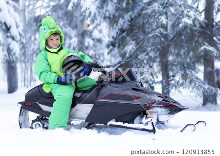 Boy sits in a green snowsuit holding a helmet on a snowmobile. 120913855