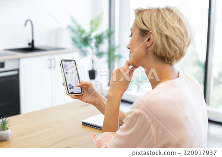 Caucasian woman in her 30s using smartphone to shop for clothing online in bright, modern kitchen. She sits at wooden table, engaged in browsing delivery website. Caucasian woman in her 30s using smartphone to shop for clothing online in bright, modern kitchen. She sits at wooden table, engaged in browsing delivery website. 120917937