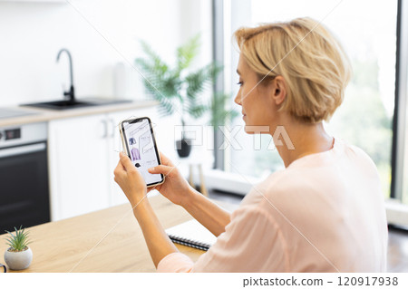 Caucasian woman in her 30s sitting at kitchen table using smartphone for online shopping. Casual attire and bright, airy kitchen setting convey relaxing and modern lifestyle. 120917938