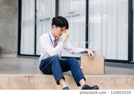 Businessman sitting outside the office with a box of belongings, reflecting on job loss. Capturing the emotional impact and reality 120920530