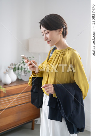 一名婦女在室內拿著包包和夾克,拿著智慧型手機 一名婦女在室內拿著包包和夾克,拿著智慧型手機 120927470