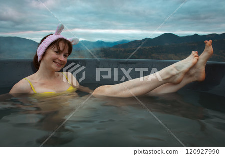 Young woman in yellow swimming suit and bunny ears in hot tub on autumn mountains landscape. Smiling girl in in outdoor jacuzzi. Happy girl in mountains resort. Healthy lifestyle. Spa relax on outdoor 120927990
