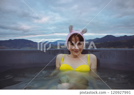 Young woman in yellow swimming suit and bunny ears in hot tub on autumn mountains landscape. Smiling girl in in outdoor jacuzzi. Happy girl in mountains resort. Healthy lifestyle. Spa relax on outdoor 120927991