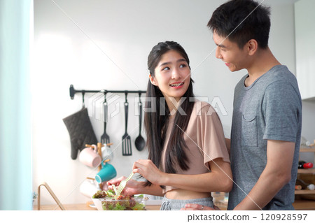 Happy cheerful Asian young couple preparing a vegetable salad in kitchen together in weekend, man holding lovely cute Maltese dog to see his girlfriend making a salad in kitchen. 120928597