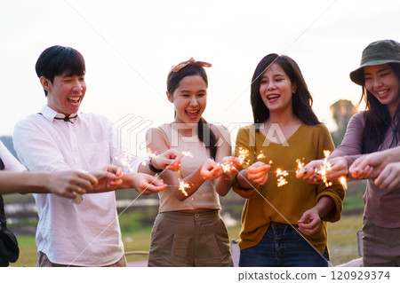 Group of Asian young men and women celebrating together with a sparkling firework in the evening. Happy men and women enjoy playing a sparklers firework in a party. 120929374