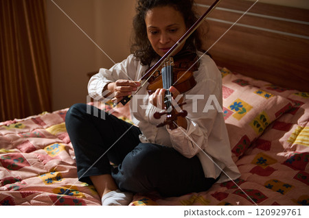 Woman playing the violin while sitting on a patterned bedspread Woman playing the violin while sitting on a patterned bedspread 120929761