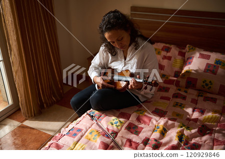 Woman practicing violin while sitting on floral bedspread in sunny room 120929846