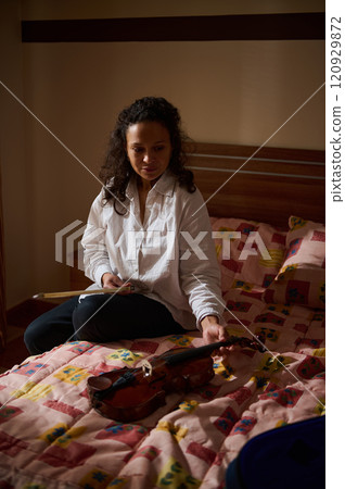 Woman sitting on bed admiring violin, basked in soft evening light Woman sitting on bed admiring violin, basked in soft evening light 120929872