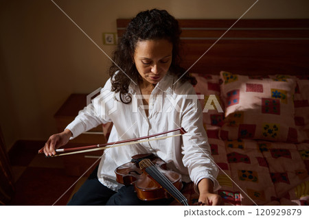 Woman practicing violin in a cozy bedroom setting with sunlight streaming 120929879