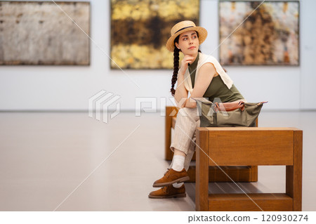 Side view of young elegantly dressed woman student in a straw hat admiring the exhibition of paintings in the modern gallery, sitting on bench. Copy space. Contemporary art and cultural education 120930274