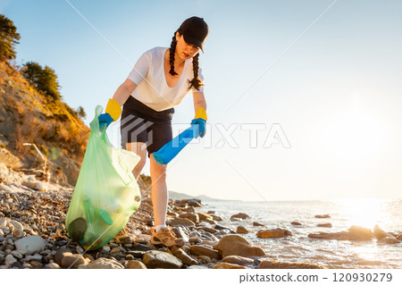 Bottom view of young Caucasian woman in rubber gloves collects garbage on pebble wild beach. Volunteer puts garbage in plastic bag. Concept of save Earth and environment disaster 120930279