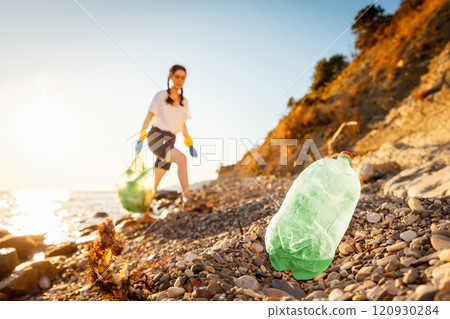 Defocused view of woman volunteer in rubber gloves holds full plastic bag of trash and walking down of pebble wild beach. Close up of plastic bottle in foreground 120930284