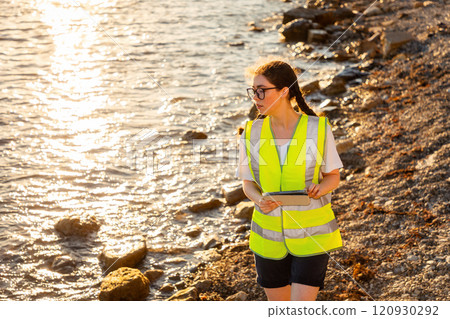 Inspection environmentalist. Young volunteer woman wearing vest and using pad for analyzing. In background is ocean, coast and sunset. Concept of environment pollution and Earth Day 120930292