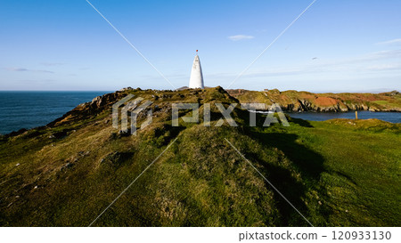 A historic lighthouse towers above the green hills overlooking the Atlantic Ocean. The bright blue sky enhances the serene atmosphere of this coastal landscape. A historic lighthouse towers above the green hills overlooking the Atlantic Ocean. The bright blue sky enhances the serene atmosphere of this coastal landscape. 120933130