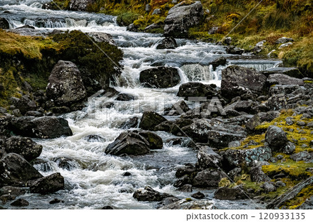 A river flows gently over smooth rocks, creating small cascading waterfalls. Surrounding grass and moss enhance the serene beauty of this natural landscape. A river flows gently over smooth rocks, creating small cascading waterfalls. Surrounding grass and moss enhance the serene beauty of this natural landscape. 120933135