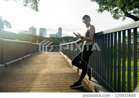 Side view of Asian sport woman lean wall railing of walkway bridge and look to right side and use mobile phone relax after exercise in public garden. Side view of Asian sport woman lean wall railing of walkway bridge and look to right side and use mobile phone relax after exercise in public garden. 120933688