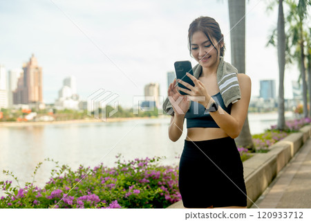Asian sport woman stand with using mobile phone and relax after exercise in public garden with high building in the city as background and soft light. 120933712