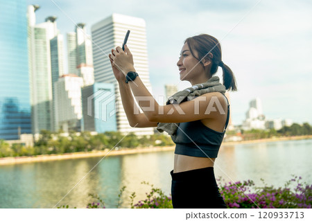 Asian sport woman use mobile phone to take photo and relax after exercise in public garden with high building of big city in background and soft light. 120933713