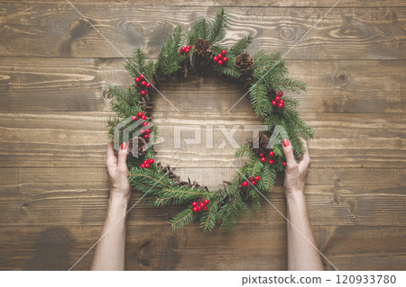 Christmas wreath made of spruce branches with holly berries on wooden board. Female hand holding wreath. Top view. Christmas wreath made of spruce branches with holly berries on wooden board. Female hand holding wreath. Top view. 120933780