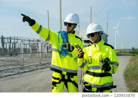 Wind turbine or windmill worker or technician man carry rope and pointing for his co-worker who use mobile phone and discuss together near small power plant of wind turbine generation. Wind turbine or windmill worker or technician man carry rope and pointing for his co-worker who use mobile phone and discuss together near small power plant of wind turbine generation. 120933870