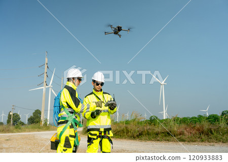 Front view wind turbine or windmill workers use drone to work in workplace field during day time and wind turbine are in the background. Front view wind turbine or windmill workers use drone to work in workplace field during day time and wind turbine are in the background. 120933883