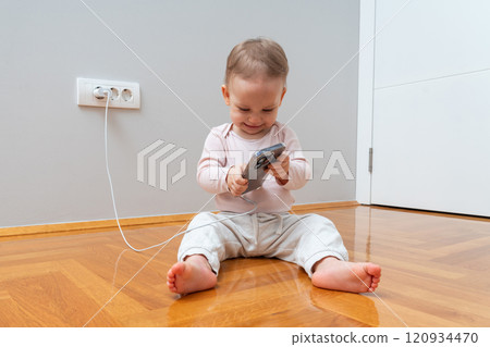 Toddler happily holding a phone and charger while sitting on a wooden floor near a wall outlet. 120934470