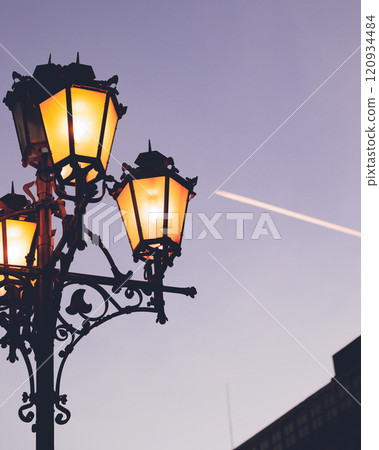 A streetlight appears at dusk with a contrail floating in it 120934484