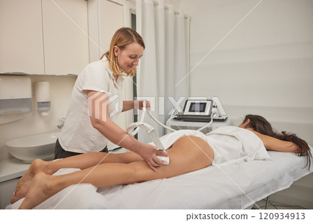 Figure correction, massage salon, non-invasive method. Young woman providing beauty services, body shaping, masseurs performing vacuum roller massage on clients, figure correction, anti-cellulite Figure correction, massage salon, non-invasive method. Young woman providing beauty services, body shaping, masseurs performing vacuum roller massage on clients, figure correction, anti-cellulite 120934913