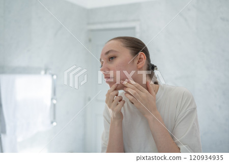 Gen z woman popping pimple on chin, stands before a mirror in bathroom. Concept of acne treatment, skincare, adult acne 120934935