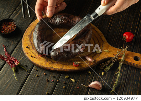 Hands carefully slice through a dark, handmade sausage on a rustic wooden board surrounded by spices and garlic, capturing the essence of culinary craftsmanship in a cozy kitchen atmosphere Hands carefully slice through a dark, handmade sausage on a rustic wooden board surrounded by spices and garlic, capturing the essence of culinary craftsmanship in a cozy kitchen atmosphere 120934977