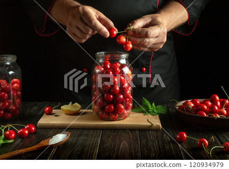 A skilled cook carefully prepares cherries for preservation, selecting the ripest fruit while surrounded by jars and fresh ingredients in a warm, rustic kitchen 120934979