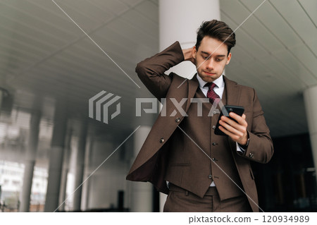 Low-angle view of worried businessman in brown suit looking surprised and concerned while checking phone standing outdoors in urban setting. Concept of business communication and technology. 120934989
