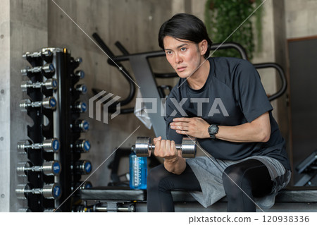 一名年輕的亞洲男子坐在健身房的訓練凳上進行肌肉訓練(手臂訓練、啞鈴、啞鈴彎舉) 一名年輕的亞洲男子坐在健身房的訓練凳上進行肌肉訓練(手臂訓練、啞鈴、啞鈴彎舉) 120938336