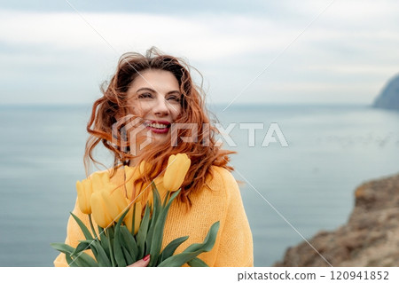 Portrait of a happy woman with hair flying in the wind against the backdrop of mountains and sea. Holding a bouquet of yellow tulips in her hands, wearing a yellow sweater 120941852