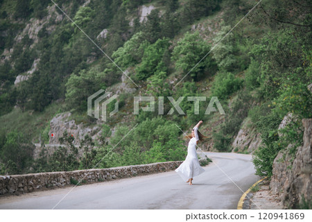 A woman is dancing on a road. The road is surrounded by trees and rocks. The woman is wearing a white dress. 120941869