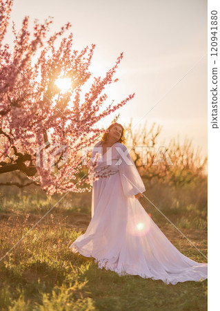 Woman blooming peach orchard. Against the backdrop of a picturesque peach orchard, a woman in a long white dress enjoys a peaceful walk in the park, surrounded by the beauty of nature. Woman blooming peach orchard. Against the backdrop of a picturesque peach orchard, a woman in a long white dress enjoys a peaceful walk in the park, surrounded by the beauty of nature. 120941880