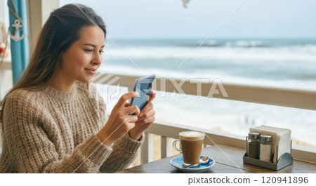 Young woman relaxing at beachside cafe with smartphone and coffee on a cozy autumn day 120941896