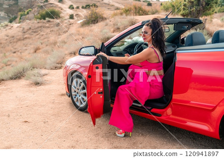 A woman in a pink outfit is sitting in a red convertible car. The car is parked on a dirt road, and the woman is enjoying the ride. 120941897