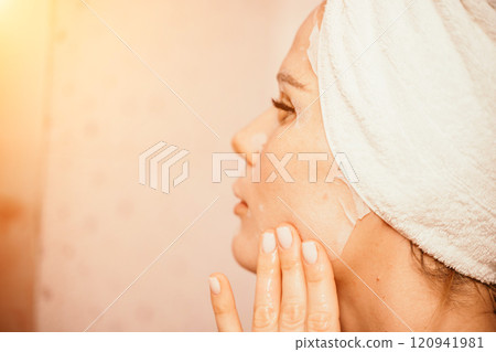 Young beautiful woman using a moisturizing facial mask after taking a bath. Pretty attractive girl in a towel on her head stands in front of a mirror in a home bathroom. Daily hygiene and skin care Young beautiful woman using a moisturizing facial mask after taking a bath. Pretty attractive girl in a towel on her head stands in front of a mirror in a home bathroom. Daily hygiene and skin care 120941981