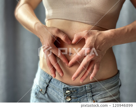 Close up of slim woman in short top and jeans forming heart shape with her hands on her bare stomach. Concept of body positivity, self love, and health awareness. Lifestyle photo. 120946899