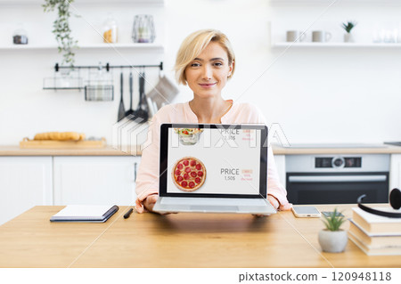 Caucasian woman showcasing online food delivery on laptop with pizza and salad. She is seated at home kitchen table, expressing happiness and convenience of digital shopping. 120948118