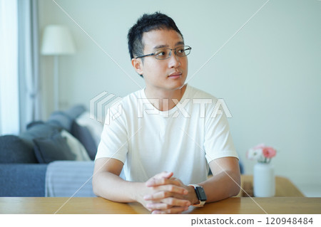 Asian man with an glasses in white t-shirt is relaxing in living room at home. A lonely man in white t-shirt portrait. 120948484