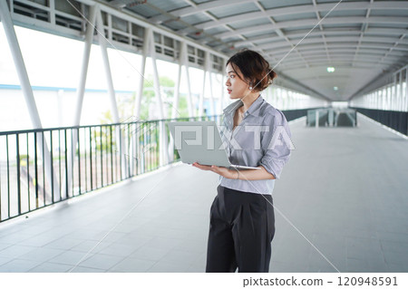Portrait of good looking Asian young businesswoman is holding and using a laptop computer while walking in the downtown. Asian businesswoman uses laptop. Portrait of good looking Asian young businesswoman is holding and using a laptop computer while walking in the downtown. Asian businesswoman uses laptop. 120948591