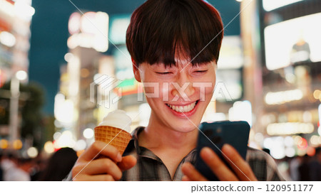 Man, phone and ice cream with travel in city, night and check notification for chat, laugh or meme in street. Person, smartphone and gelato on metro sidewalk, smile and app for social media in Japan 120951177