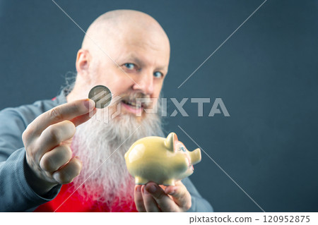 an adult bald man with a large gray beard puts a coin in a piggy bank 120952875