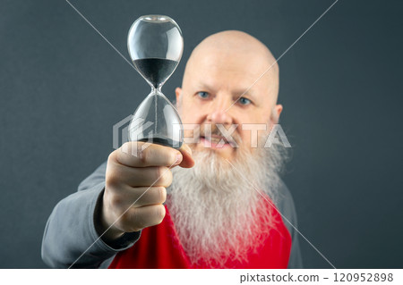 bald man with a large white beard holds an hourglass in his hands. concept of fluidity of life time 120952898