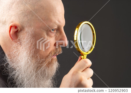 serious man with a long gray beard looks through a magnifying glass 120952903